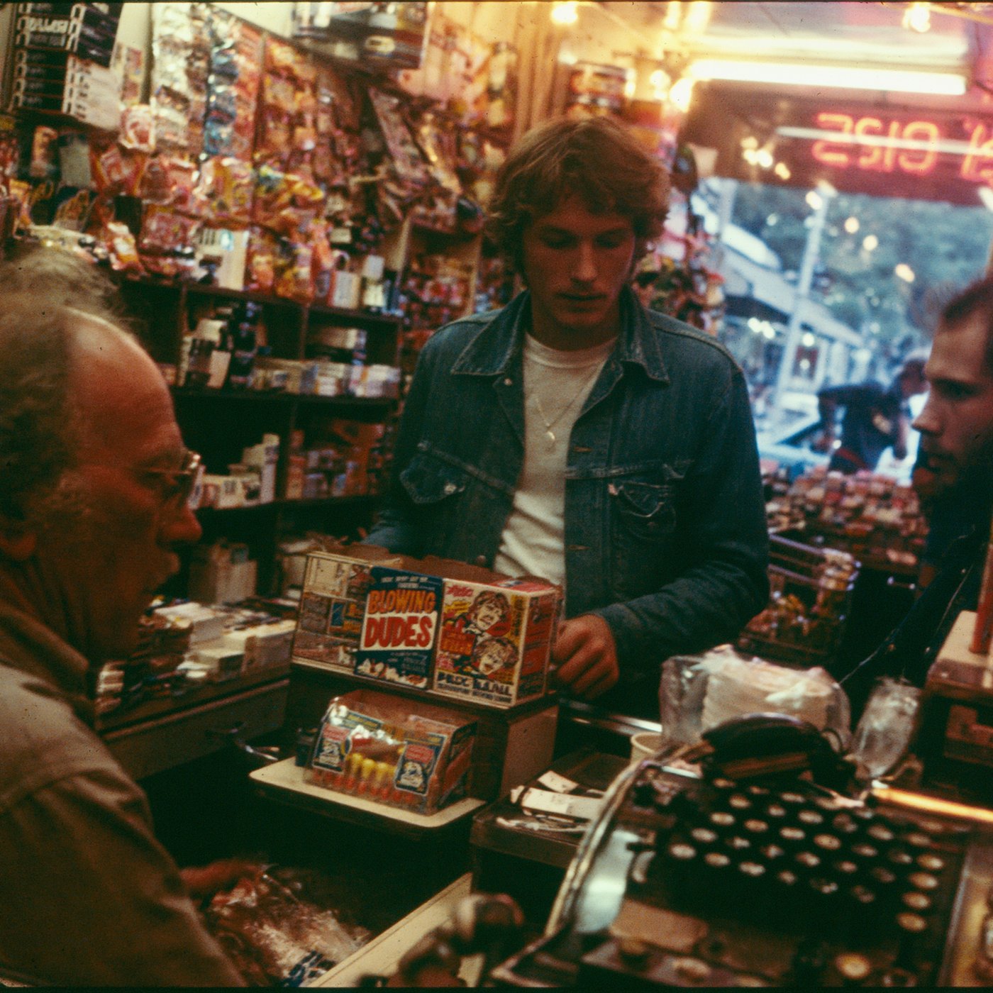 A young dude in a denim jacket at a 1970s corner store counter buying from a Blowing Dudes display box
